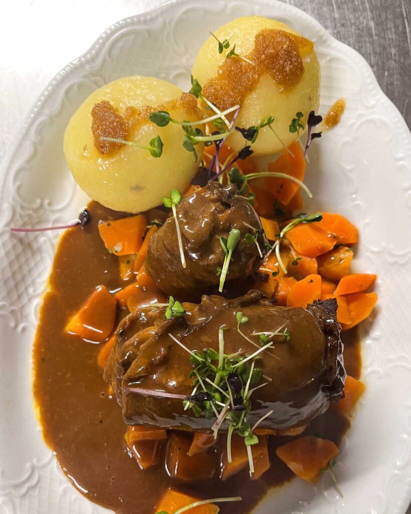 Zarte Rinderroulade in dunkler Soße mit frischen Karotten und zwei Klößen, angerichtet auf einem weißen Teller im Brauereigasthof Werneck.